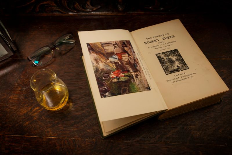 A book open displaying the poetry of Robert Burns beside a glass of whisky and pair of reading glasses