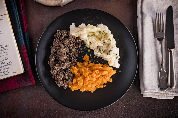 A plate of haggis, neeps and mashed potatoes on a table beside a knife, fork and napkin and a book